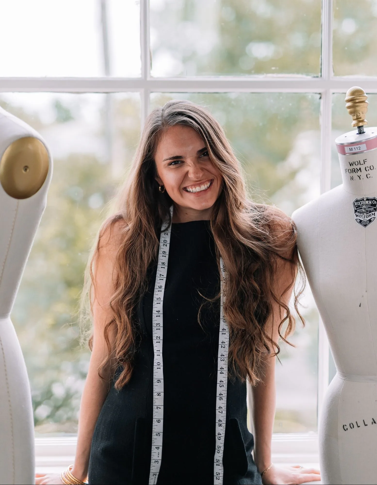 Samantha Covey in the atelier Samantha Covey smiling between dress forms in front of an atelier window.